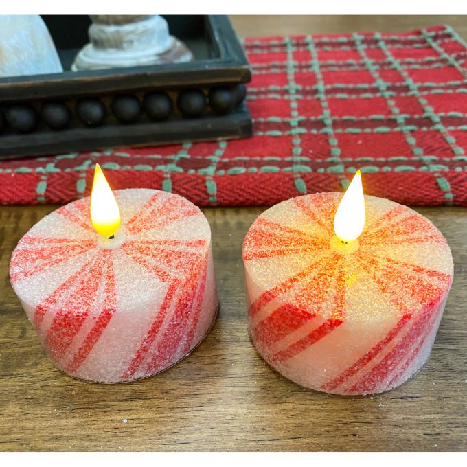 Two striped candles with flames on a wooden surface with a red checkered cloth in the background. Short Peppermint Votive LED Timer Candles - Set of Two available at Quilted Cabin Home Decor.