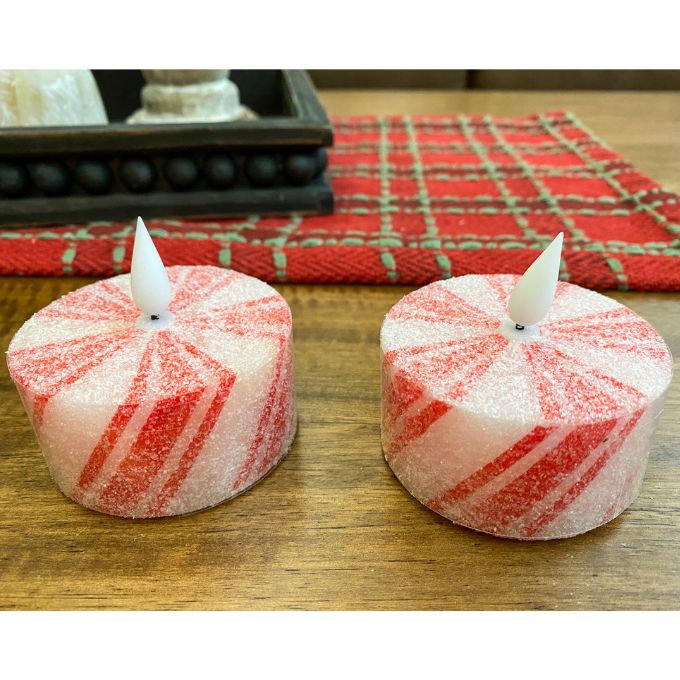Two red and white striped candles on a wooden surface with a plaid cloth in the background. Short Peppermint Votive LED Timer Candles - Set of Two available at Quilted Cabin Home Decor.