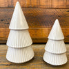 Two white ceramic Christmas trees on a wooden surface with a wooden background. Tiered Distressed Tan Ceramic Trees - Two Sizes available at Quilted Cabin Home Decor.