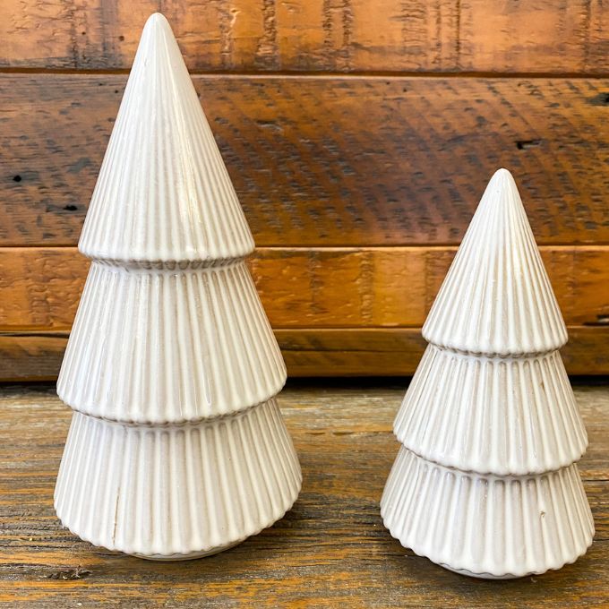 Two white ceramic Christmas trees on a wooden surface with a wooden background. Tiered Distressed Tan Ceramic Trees - Two Sizes available at Quilted Cabin Home Decor.