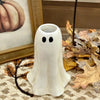 A small white ghost-shaped candle holder with black eyes sits on a woven rug surrounded by dried autumn leaves and a framed pumpkin print. Available in Airdrie at Quilted Cabin 