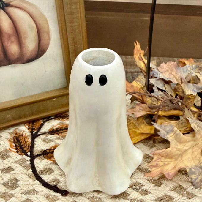 A small white ghost-shaped candle holder with black eyes sits on a woven rug surrounded by dried autumn leaves and a framed pumpkin print. Available in Airdrie at Quilted Cabin 