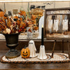 Halloween-themed tabletop decor set featuring a white ghost-shaped candle holder, a ceramic pumpkin lantern, and a ghost-shaped candle holder, alongside a large urn with autumn leaves and a rustic framed painting of ghosts.  Available at Quilted Cabin in Airdrie