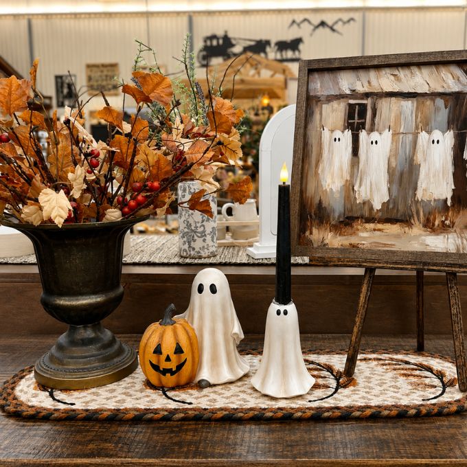 Halloween-themed tabletop decor set featuring a white ghost-shaped candle holder, a ceramic pumpkin lantern, and a ghost-shaped candle holder, alongside a large urn with autumn leaves and a rustic framed painting of ghosts.  Available at Quilted Cabin in Airdrie