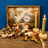 A framed autumn landscape print showing a white barn among fall trees, styled on a rustic wooden surface with scattered leaves, acorns, a small owl figurine, and two lit green candles against a deep blue background. All items are available at Quilted Cain in Airdrie