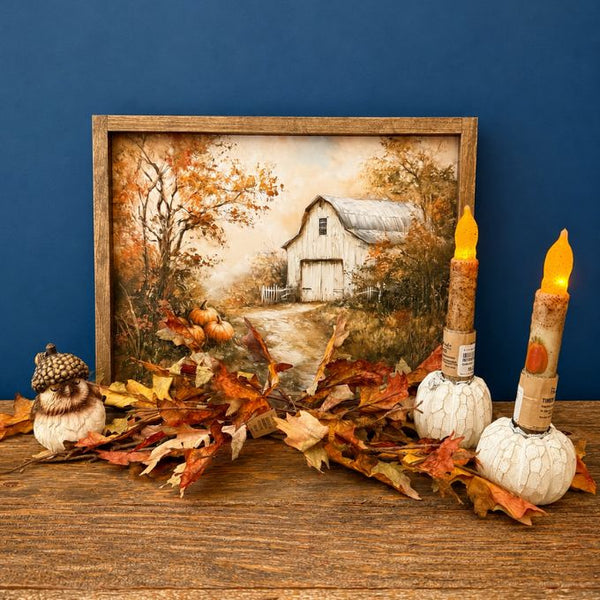A framed autumn landscape print showing a white barn among orange and brown trees, styled on a wooden surface with scattered fall leaves, a small owl figurine, and two white candle holders with lit flameless candles against a dark blue background. All items available at Quilted Cabin in Airdrie