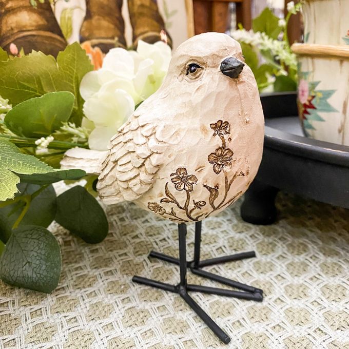 Decorative bird figurine with floral patterns on a stand, surrounded by greenery and a candle. Engraved Floral Bird Figurine available at Quilted Cabin Home Decor.