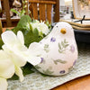 Decorative ceramic bird with floral patterns on a table with flowers and a placemat. Painted Floral Porcelain Bird available at Quilted Cabin Home Decor.