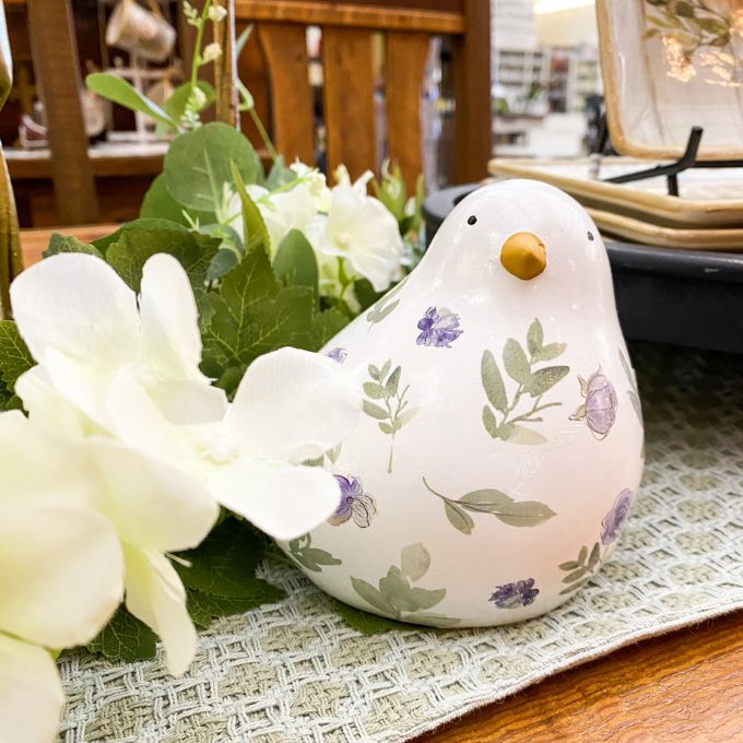 Decorative ceramic bird with floral patterns on a table with flowers and a placemat. Painted Floral Porcelain Bird available at Quilted Cabin Home Decor.