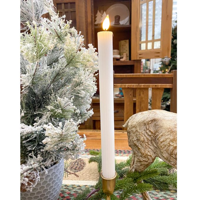 Tall white candle in a gold holder with a Christmas tree and decorative elements in the background. White Ridged LED Timer Taper Candles - Two Sizes available at Quilted Cabin Home Decor.