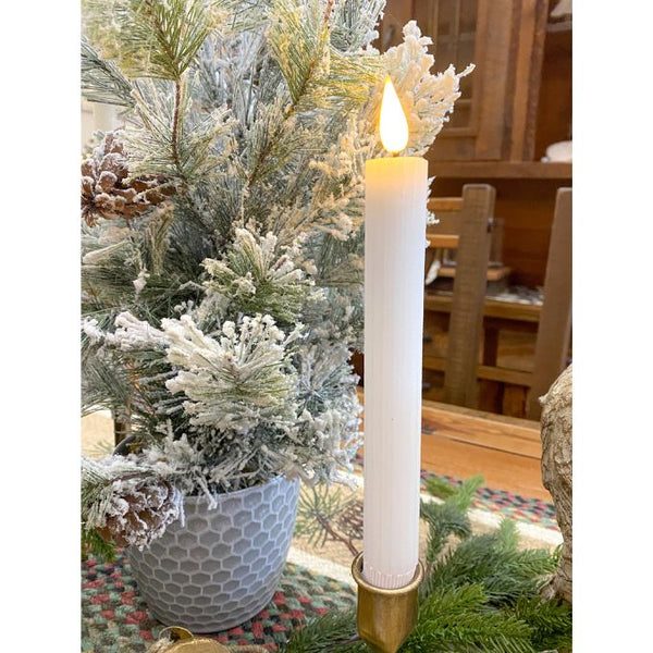 Decorative candle in a gold holder with a Christmas tree in the background. White Ridged LED Timer Taper Candles - Two Sizes available at Quilted Cabin Home Decor.