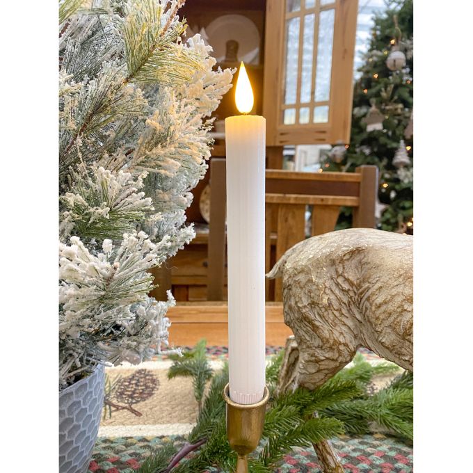 Decorative candle in a gold holder with Christmas trees and a wooden bench in the background. White Ridged LED Timer Taper Candles - Two Sizes available at Quilted Cabin Home Decor.