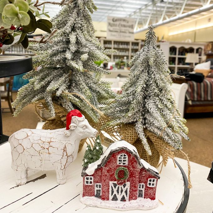 Decorative Christmas scene with a cow figurine, small red barn, and trees in a store setting. Snowy Red Barn Figurine available at Quilted Cabin Home Decor.