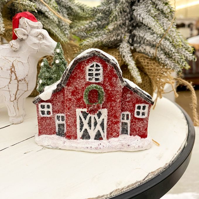 Decorative red barn figurine with snow on a white surface, Christmas tree in the background. Snowy Red Barn Figurine available at Quilted Cabin Home Decor.