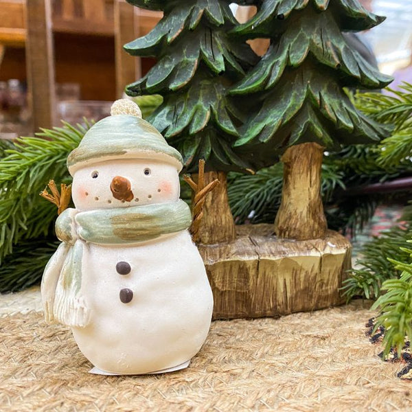 Decorative snowman figurine with a green hat and scarf in front of a Christmas tree. Green Hat Snowman Figurine available at Quilted Cabin Home Decor.