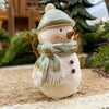 Decorative snowman figurine with a green hat and scarf on a textured surface with blurred greenery in the background. Green Hat Snowman Figurine available at Quilted Cabin Home Decor.