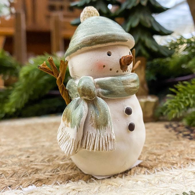 Decorative snowman figurine with a green hat and scarf on a textured surface with blurred greenery in the background. Green Hat Snowman Figurine available at Quilted Cabin Home Decor.