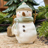 Decorative snowman figurine with a green hat and scarf, standing on a textured surface with greenery in the background. Green Hat Snowman Figurine available at Quilted Cabin Home Decor.