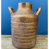 Gold ceramic vase with handles on a wooden surface against a blue background. Gold Metal Milk Can Vase available at Quilted Cabin Home Decor.