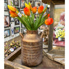 Decorative vase with orange tulips in a store setting with framed pictures on the wall. Gold Metal Milk Can Vase available at Quilted Cabin Home Decor.