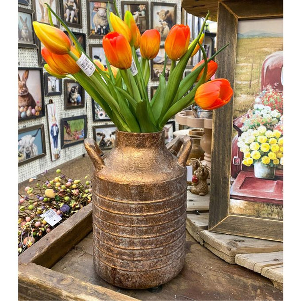 Decorative vase with orange tulips in a store setting with framed pictures on the wall. Gold Metal Milk Can Vase available at Quilted Cabin Home Decor.