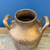 Rustic gold vase with handles on a wooden surface against a blue background. Gold Metal Milk Can Vase available at Quilted Cabin Home Decor.