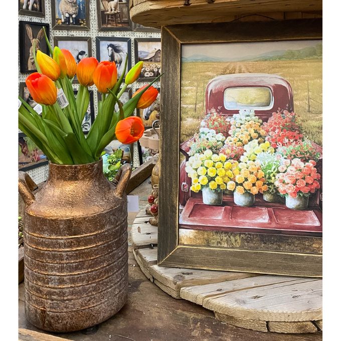 Decorative vase with orange tulips next to a framed picture of a truck with flowers on a wooden surface. Gold Metal Milk Can Vase available at Quilted Cabin Home Decor.