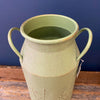 Green ceramic vase with handles on a wooden surface against a blue wall. Embossed Metal Milk Cans - Two Colours available at Quilted Cabin Home Decor.