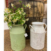 Two metal jugs, one green and one white, with floral decorations, next to a wooden barrel. Embossed Metal Milk Cans - Two Colours available at Quilted Cabin Home Decor.
