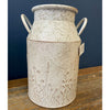 Decorative ceramic milk can with floral patterns on a wooden surface against a blue wall. Embossed Metal Milk Cans - Two Colours available at Quilted Cabin Home Decor.