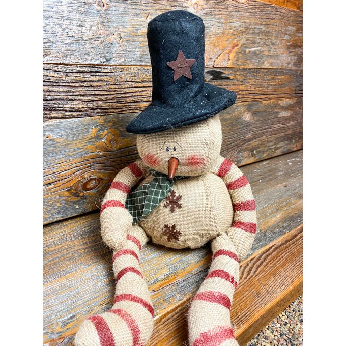 A burlap snowman with long limbs with red candy cane style stripes. It has a tall black hat and  a a green fabric scary. It is available at Quilted Cabin Home Decor.