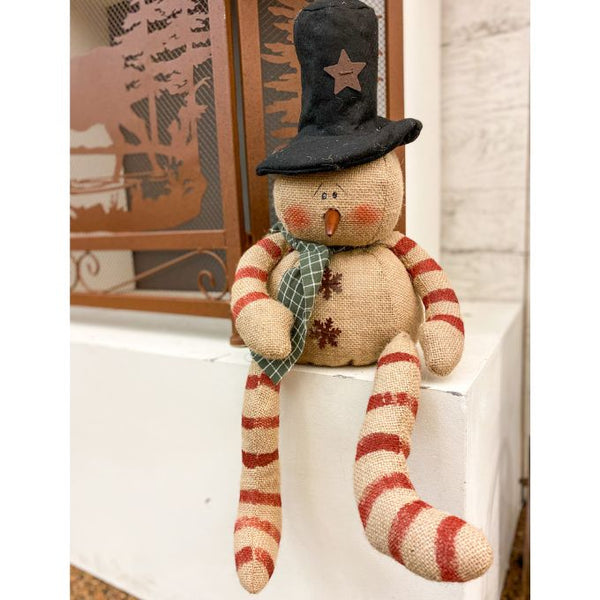 A burlap snowman with long limbs with red candy cane style stripes. It has a tall black hat and  a a green fabric scary. It is available at Quilted Cabin Home Decor.