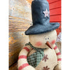 A burlap snowman with long limbs with red candy cane style stripes. It has a tall black hat and  a a green fabric scary. It is available at Quilted Cabin Home Decor.