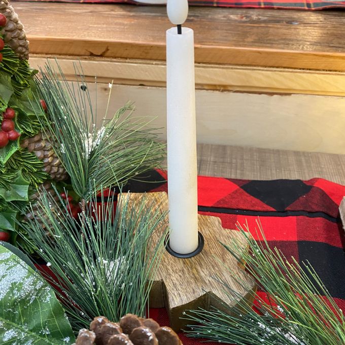 White candle in a wooden tree shaped holder with greenery and a red checkered fabric in the background. Wooden Tree Taper Holder available at Quilted Cabin Home Decor.