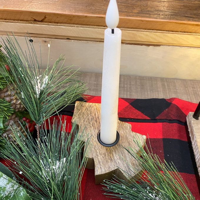 White candle in a wooden holder on a red and black checkered surface with greenery. Wooden Tree Taper Holder available at Quilted Cabin Home Decor.