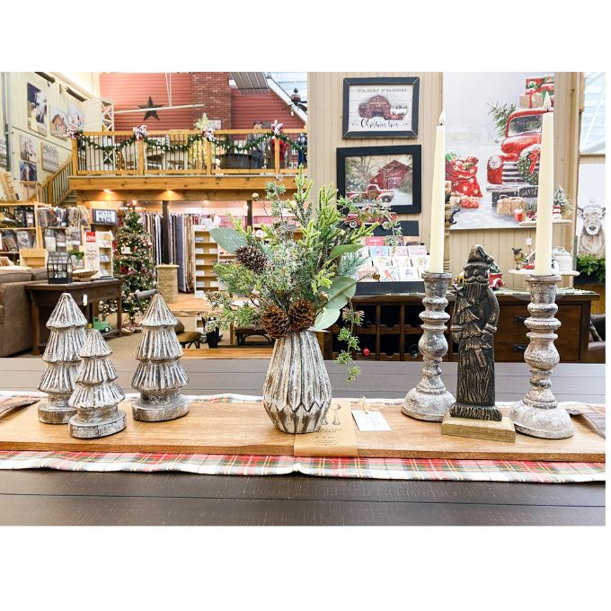 Decorative items including a vase with greenery, candle holders, and Christmas trees on a table in a store setting. Tarnished Santa Figurine available at Quilted Cabin Home Decor.