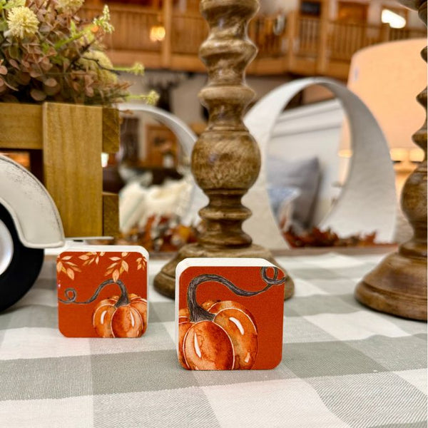 Two square decorative coasters with a fall pumpkin design, placed on a gray and white checkered tablecloth in a rustic indoor setting. Available at Quilted Cabin in Airdrie