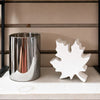 A polished silver cylindrical container and a white maple leaf-shaped decorative object displayed on a light fabric surface against a beige paneled background. Maple leaf cutout available at Quilted Cabin in Airdrie