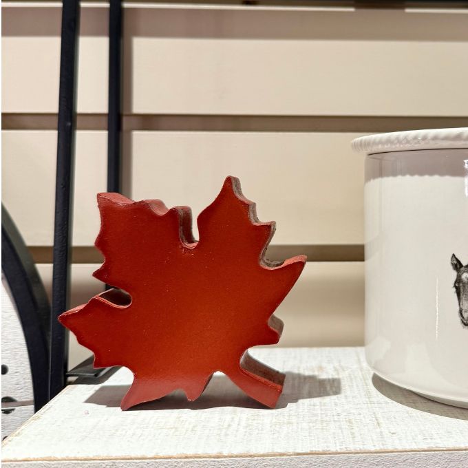 A red maple leaf-shaped decorative object displayed on a light textured surface next to a white ceramic container with a horse illustration.  Maple Leaf Cutout available at Quilted Cabin in Airdrie