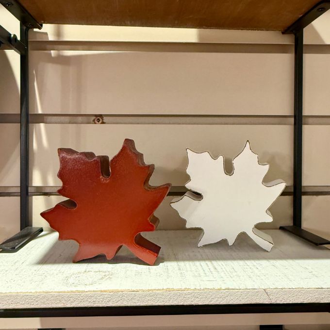 Two decorative maple leaf wall art pieces displayed on a shelf, one in reddish-brown and one in white, set against a beige slatwall background.  Maple Leaf cutout available at Quilted Cabin in Airdrie