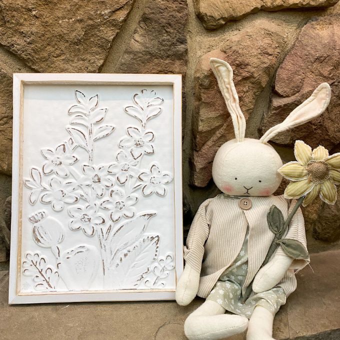 Stuffed bunny toy with a flower against a stone wall, next to a framed floral artwork. Embossed Whitewashed Flower Frame available at Quilted Cabin Home Decor.
