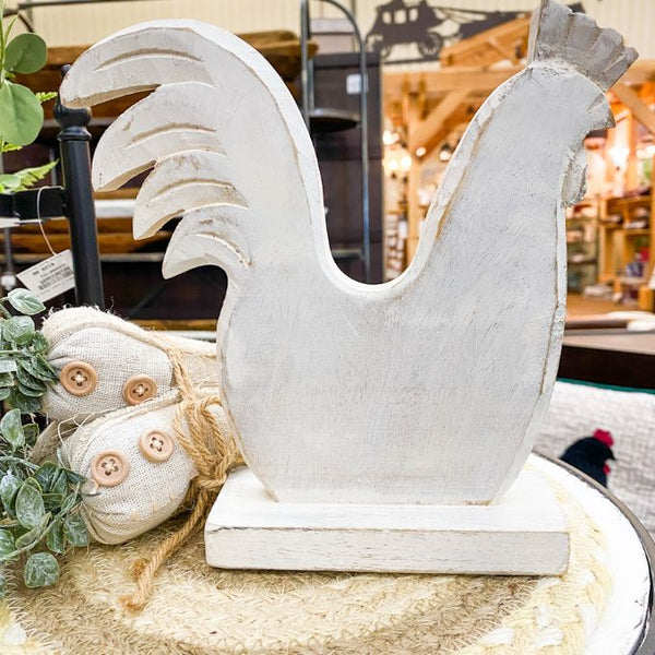 Decorative white hen on a table with plants and a blurred background. Whitewashed Wooden Hen on Stand available at Quilted Cabin  Home Decor. 
