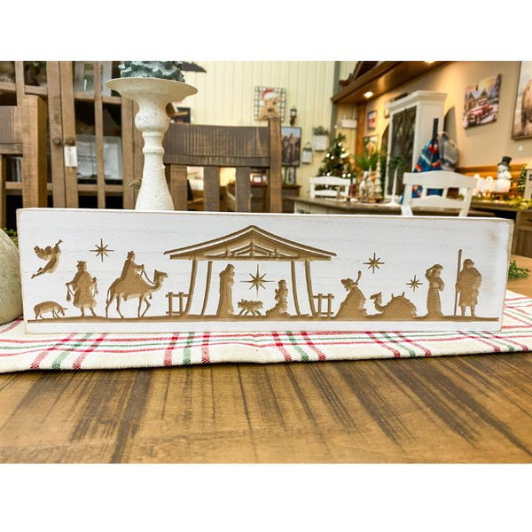 Decorative nativity scene box on a table with a plaid cloth in a home setting. Distressed White Carved Nativity Block Sign available at Quilted Cabin Home Decor.