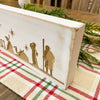 Wooden block with nativity scene design on a plaid tablecloth. Distressed White Carved Nativity Block Sign available at Quilted Cabin Home Decor.