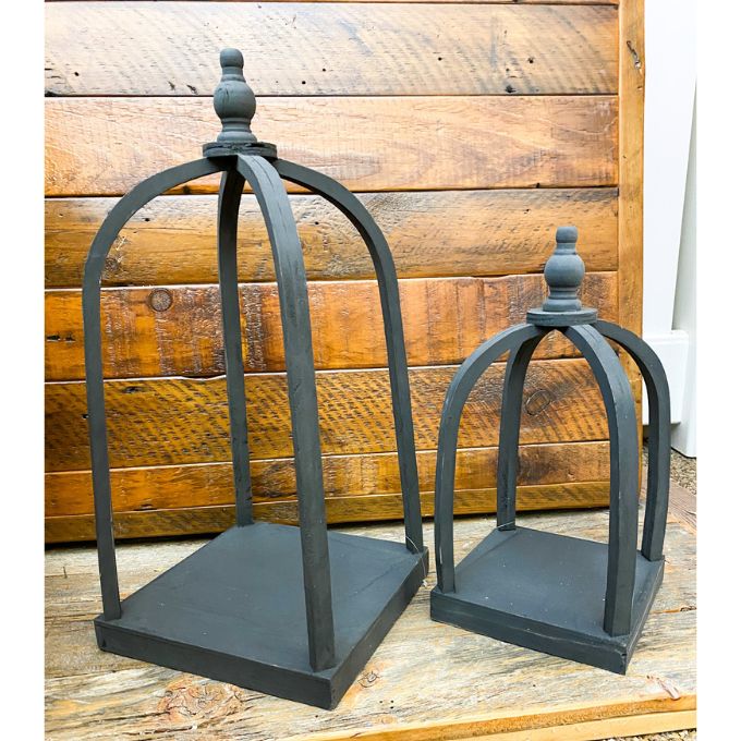 Two black wood lanterns on a wooden surface with a wooden wall background. Black Curved Top Lantern - Two Sizes available at Quilted Cabin Home Decor.