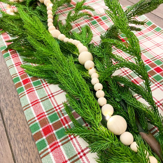 A light wood beaded garland with lights throughout is shown as part of Christmas decor. Available at Quilted Cabin Home Decor.