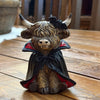 A small decorative figurine of a shaggy Highland cow dressed as a vampire, sitting on a wooden surface. The figurine features a black cape with red lining, a black bow on its head, and a metallic gold-brown textured coat. Available at Quilted Cabin in Airdrie