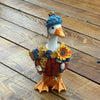 A decorative goose figurine wearing a blue knit beanie, green and brown knit sweater, and matching scarf, holding a small orange pumpkin, set on a wooden plank surface.  Available at Quilted Cabin in Airdrie
