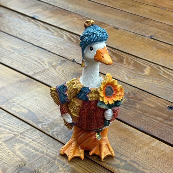A decorative goose figurine wearing a blue knit beanie, green and brown knit sweater, and matching scarf, holding a small orange pumpkin, set on a wooden plank surface.  Available at Quilted Cabin in Airdrie