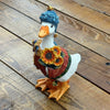 A decorative goose figurine wearing a blue knit beanie, green and brown knit sweater, and matching scarf, holding a small orange pumpkin, set on a wooden plank surface.  Available at Quilted Cabin in Airdrie